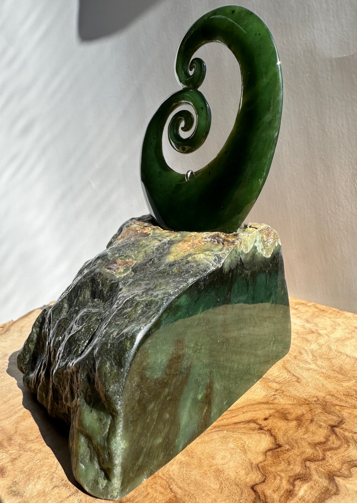 A green polished stone sculpture featuring a curved spiral motif mounted on a rough, natural stone base, displayed on a wooden surface against a plain light background.
