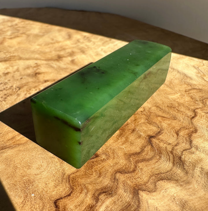 A polished green jade rectangular block with natural dark inclusions, resting on a textured wooden surface under direct sunlight.