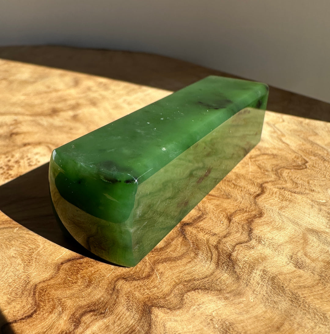 A polished green jade block resting on a textured, wavy wood surface with direct sunlight casting a shadow.