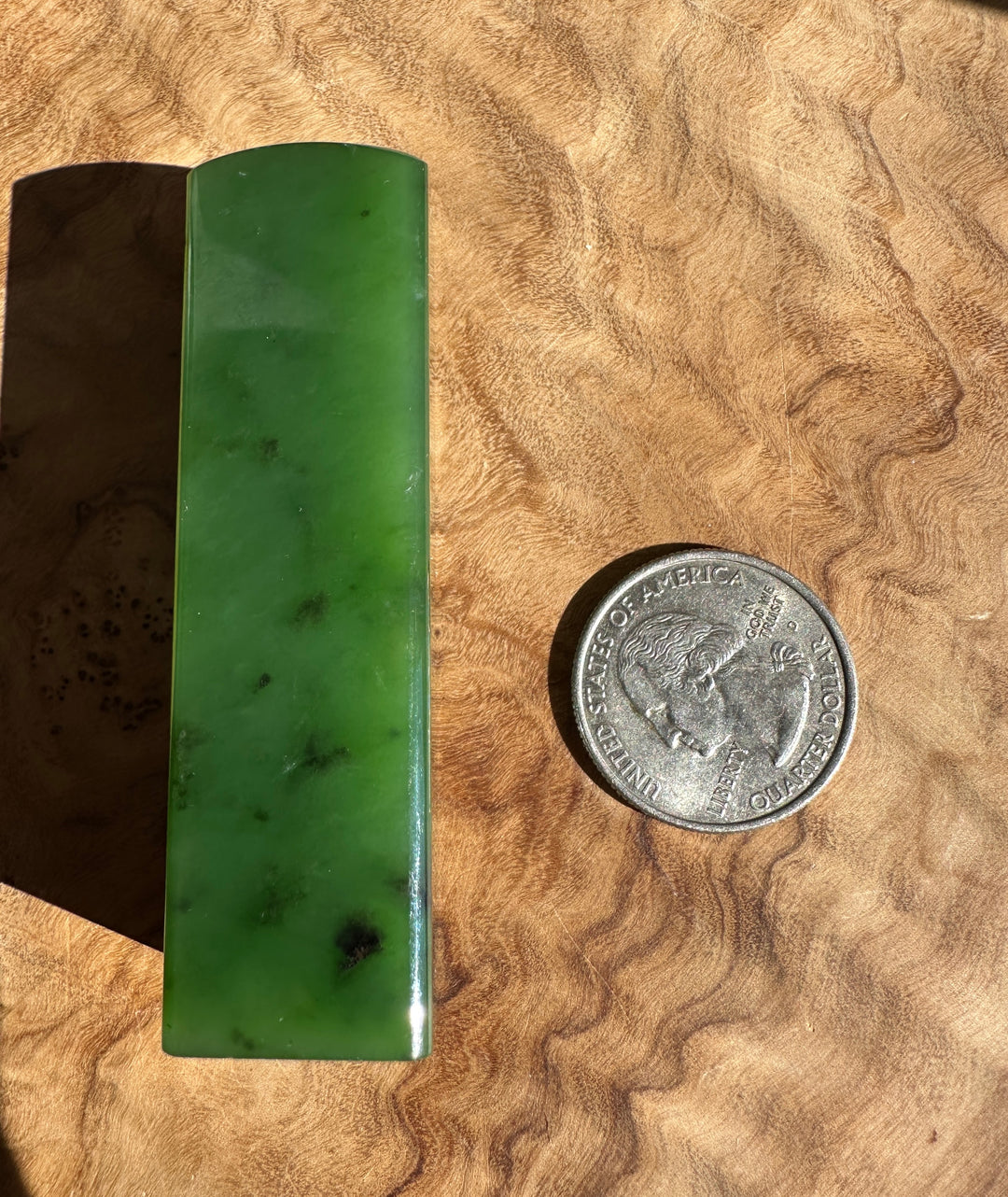 A rectangular green jade block with a curved top, shown next to a US quarter coin for scale on a wavy wood surface.