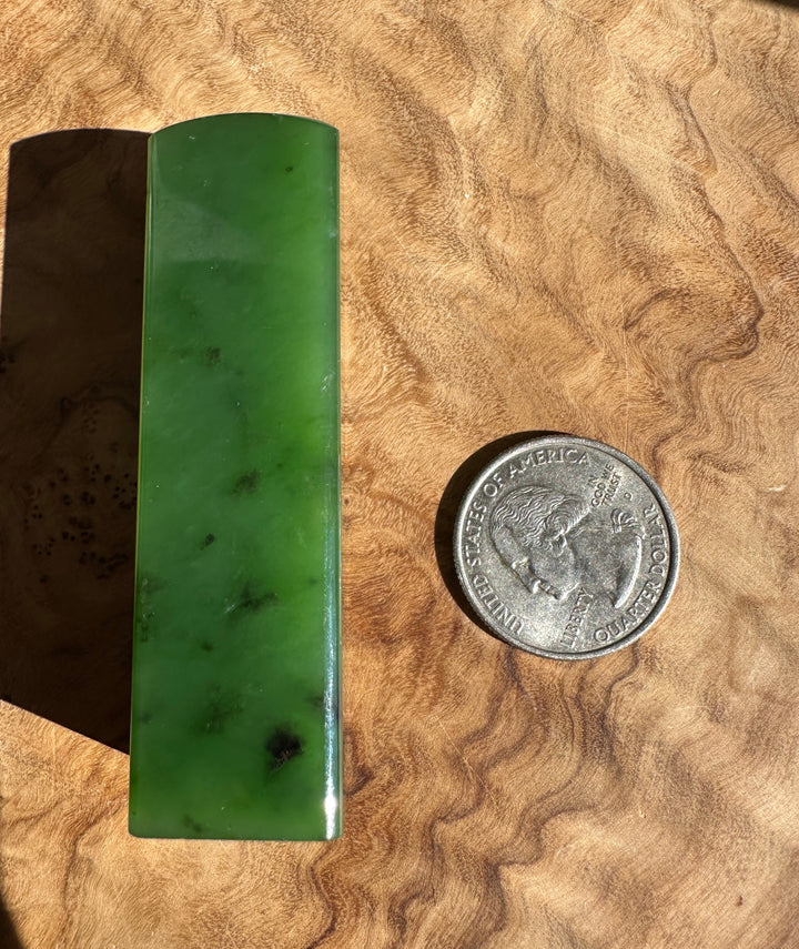 A rectangular green jade block with a curved top, shown next to a US quarter coin for scale on a wavy wood surface.