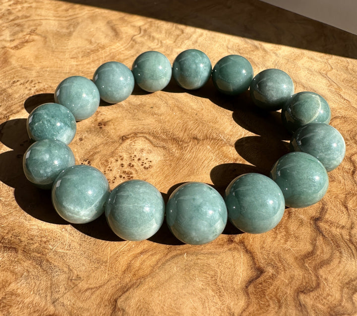 A beaded bracelet made of polished round blue/green gemstone beads, arranged in a circle on a textured wooden surface with direct sunlight casting shadows.