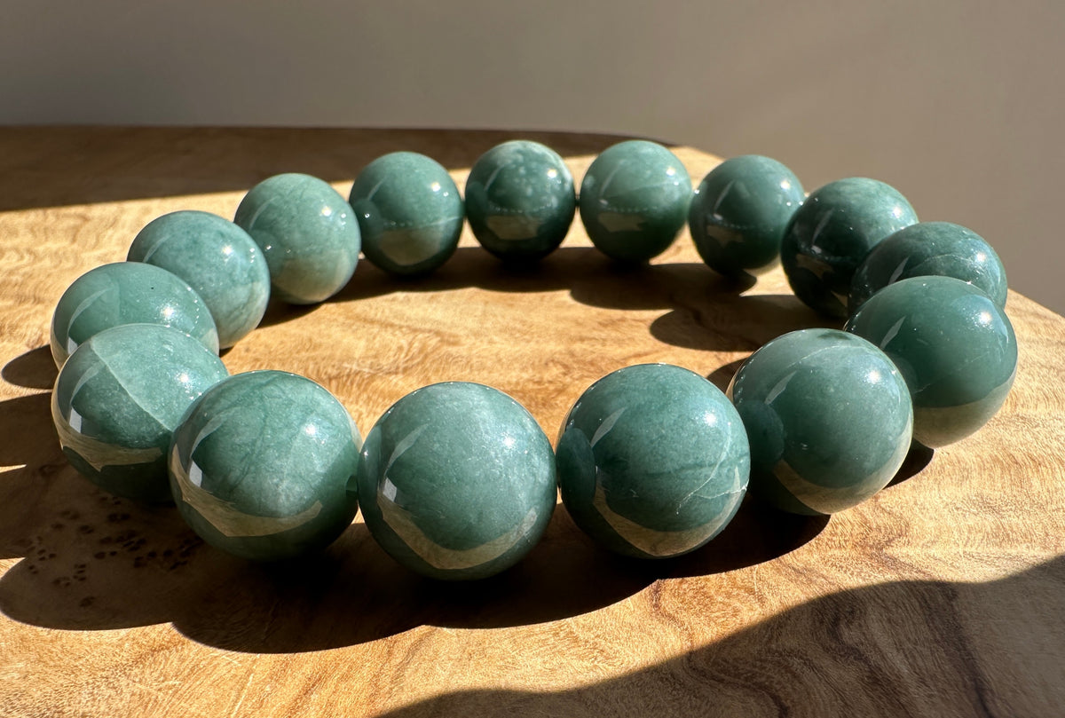 A beaded bracelet made of polished round blue/green stone beads, arranged in a circle on a wooden surface with natural sunlight casting shadows.