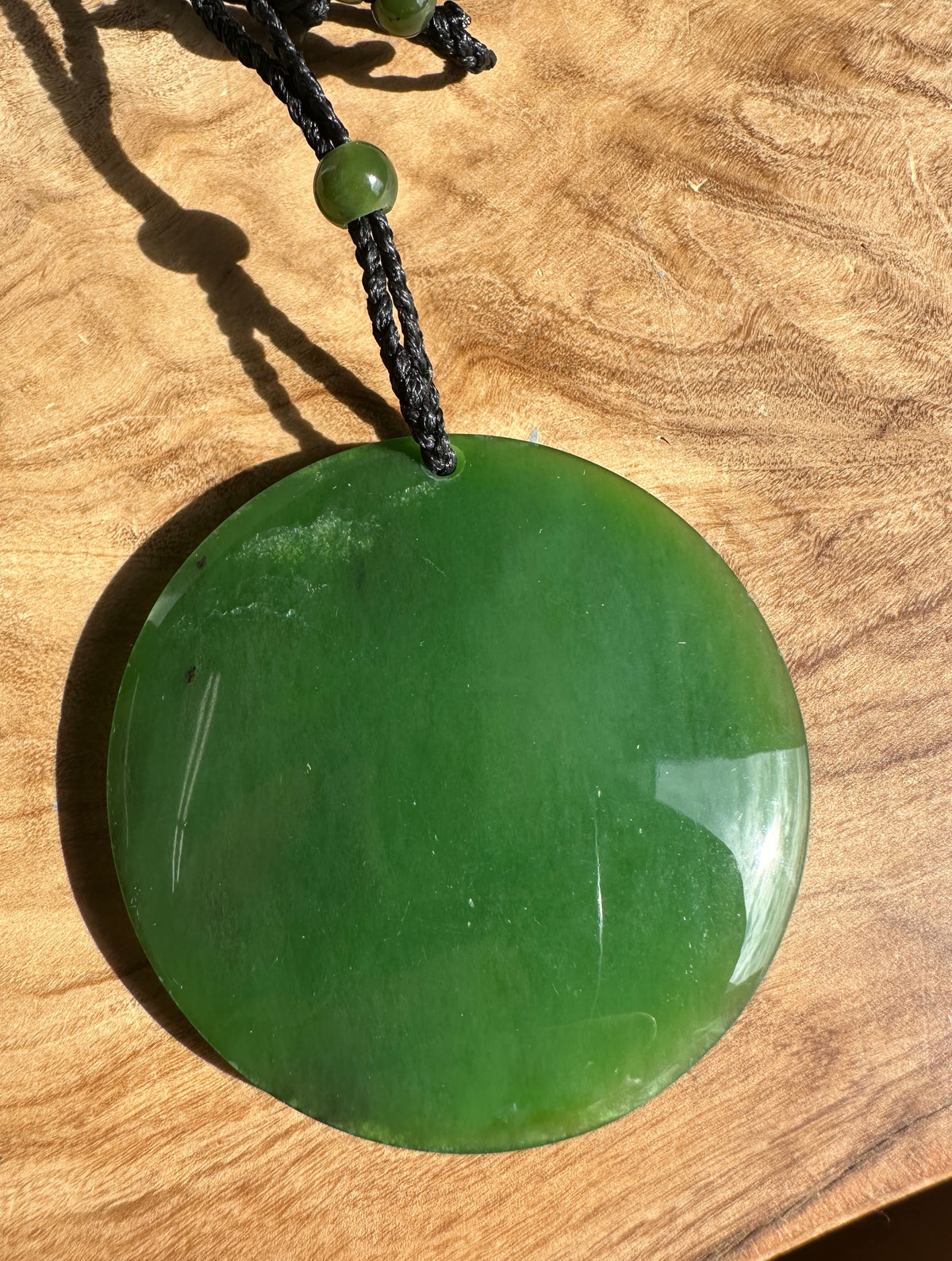 A round green jade pendant necklace with a black cord and a small green bead, displayed on a wooden surface.