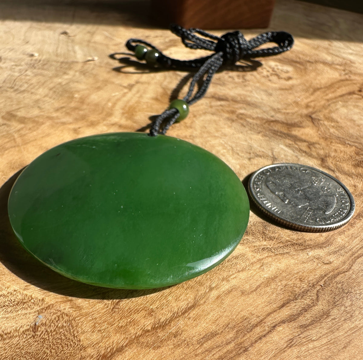 A polished green jade pendant necklace with a black braided cord and a small green bead, displayed on a wooden surface next to a coin for scale.