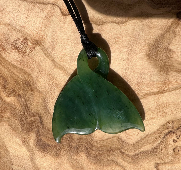 Green jade whale tail pendant with layered fins on a black cord against a wood background