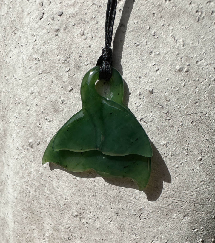 Close up of green jade whale tail pendant with a top loop on a black cord against a textured stone wall
