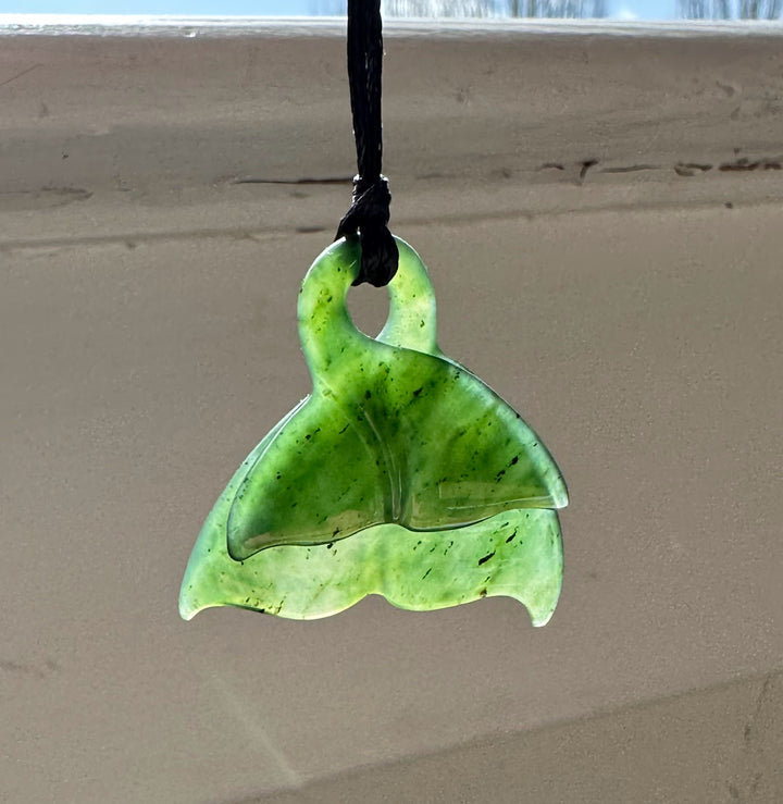Green jade whale tail pendant with a top loop on a black cord against a neutral background.