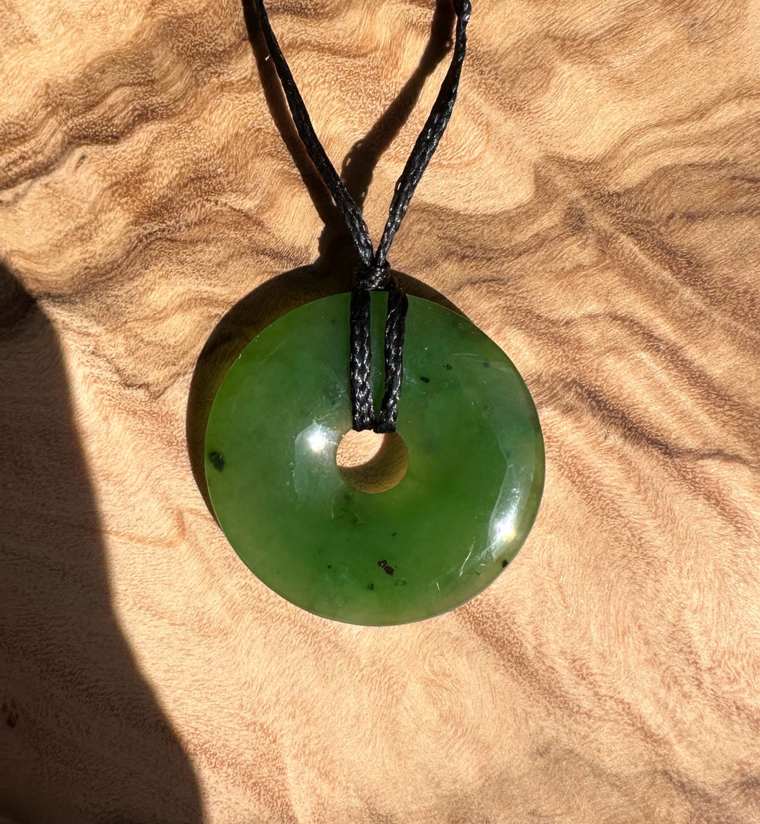 Green Canadian Jade 25mm donut pendant on a black cord resting on tan fabric.