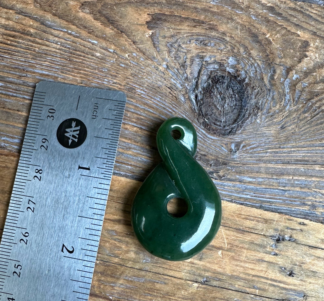 Dark green jade infinity twist pendant with a hole for stringing placed on wood beside a ruler for scale
