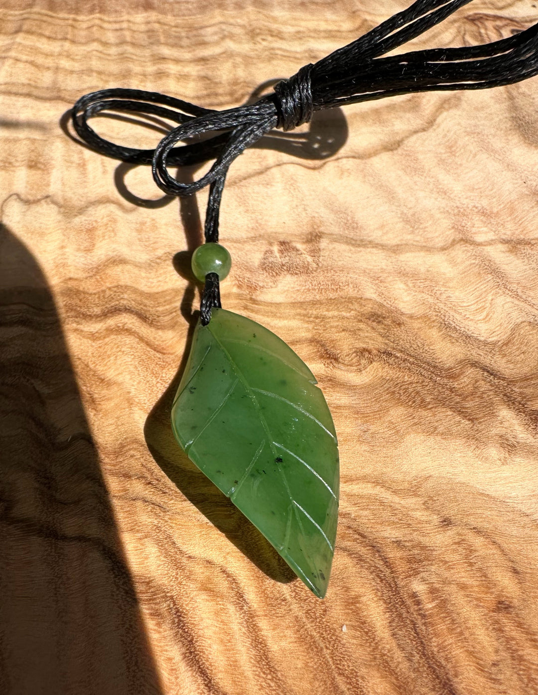 Pale green jade leaf pendant on a black cord with a round bead against wood background