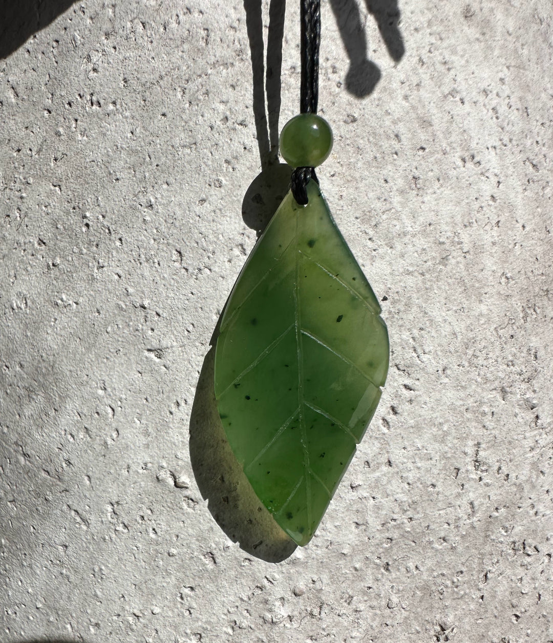 Green jade leaf pendant with vein pattern on black cord against a light textured surface