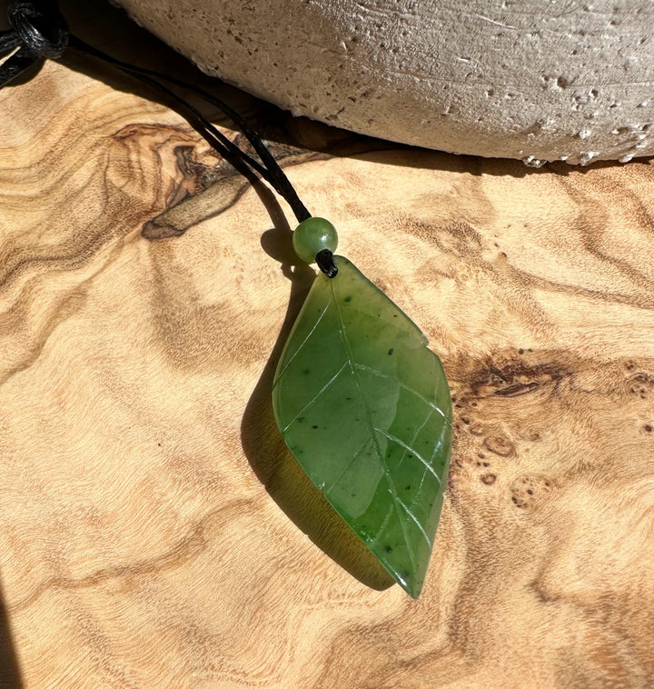 Green jade leaf pendant on black cord with a small bead, placed on wood background.