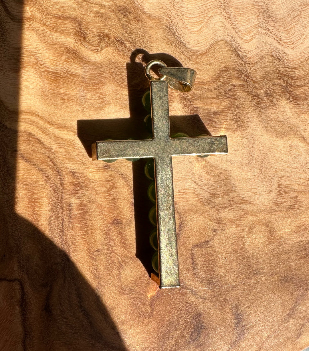 Jade bead cross pendant resting on wood, showing metal frame and bail