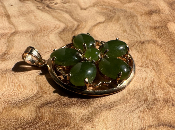 Vintage jade flower pendant resting on a wood surface showing a gold circular frame and five oval green jade petals around a central bead