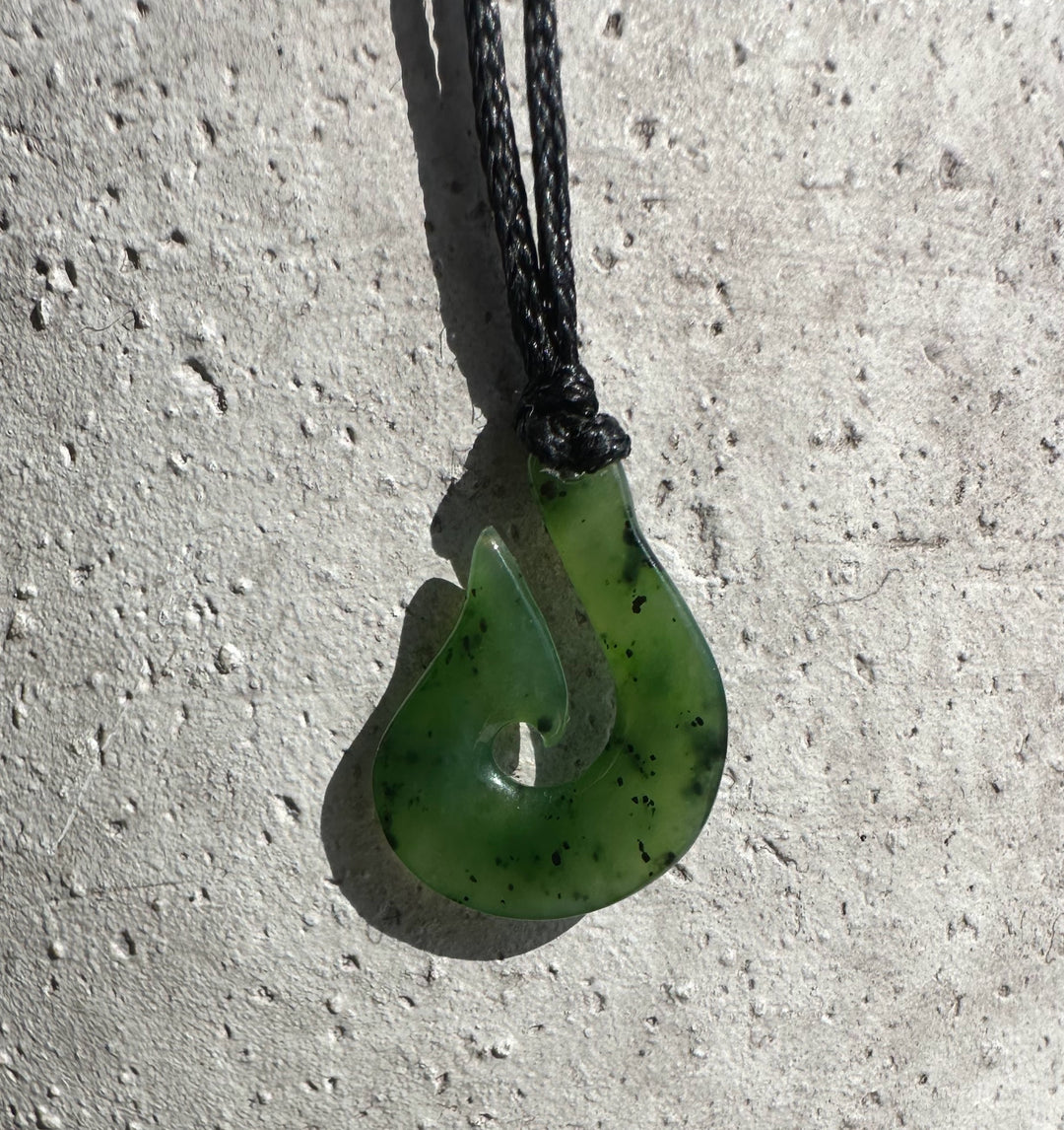 Small green jade fish hook pendant on a black braided cord against a light textured wall.