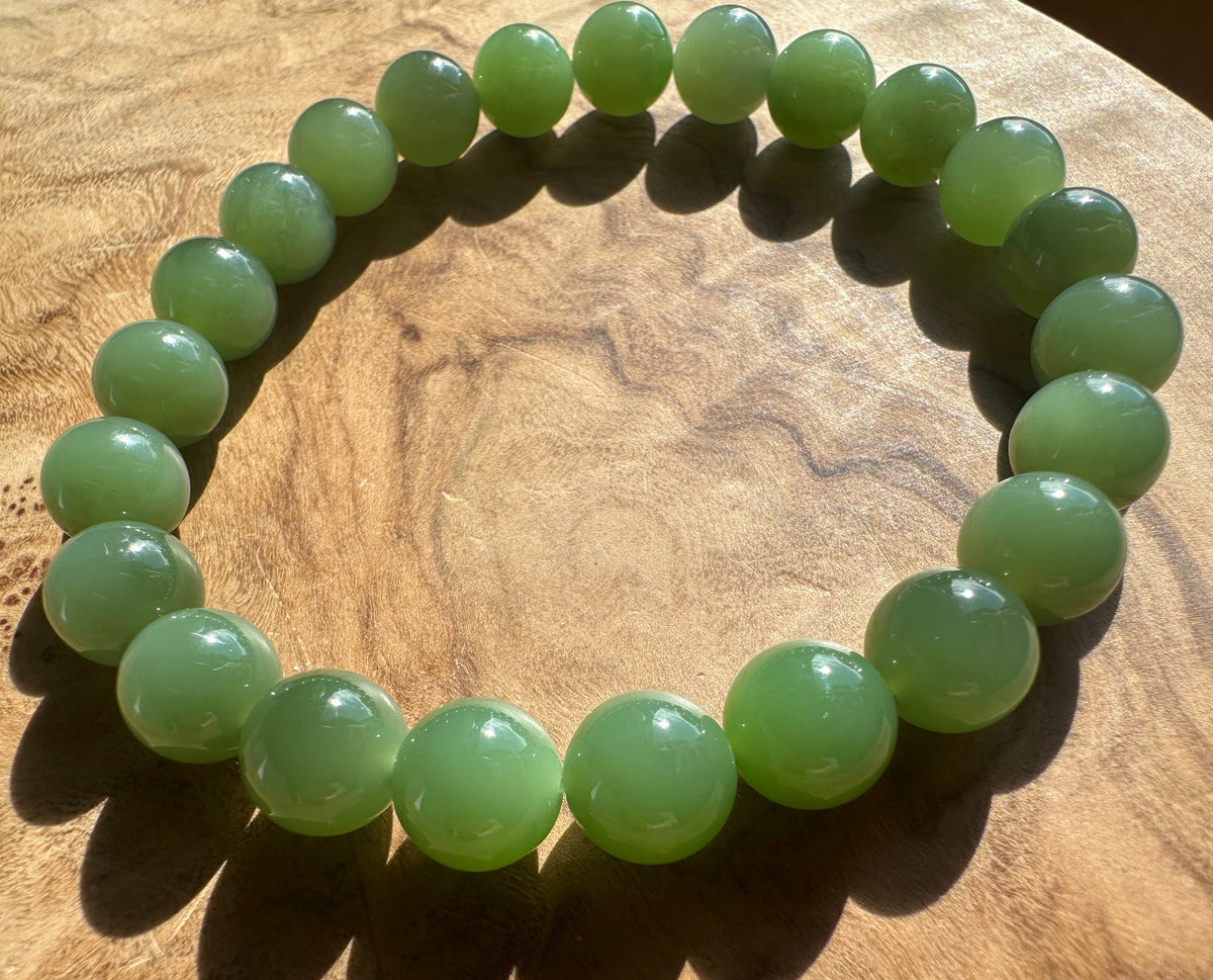 A beaded stretch bracelet made of smooth, polished light green jade round beads, displayed on a textured wooden surface.