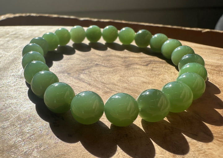 A beaded stretch bracelet made of smooth, polished light green jade-like round beads, displayed on a wooden surface with natural sunlight casting shadows.