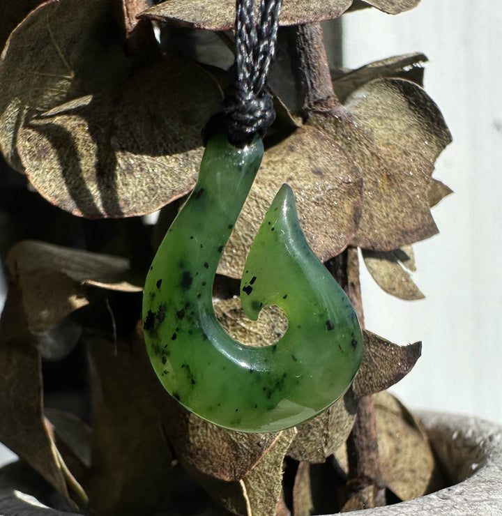 Green jade fish hook pendant with dark specks on a black cord against a natural wood background.