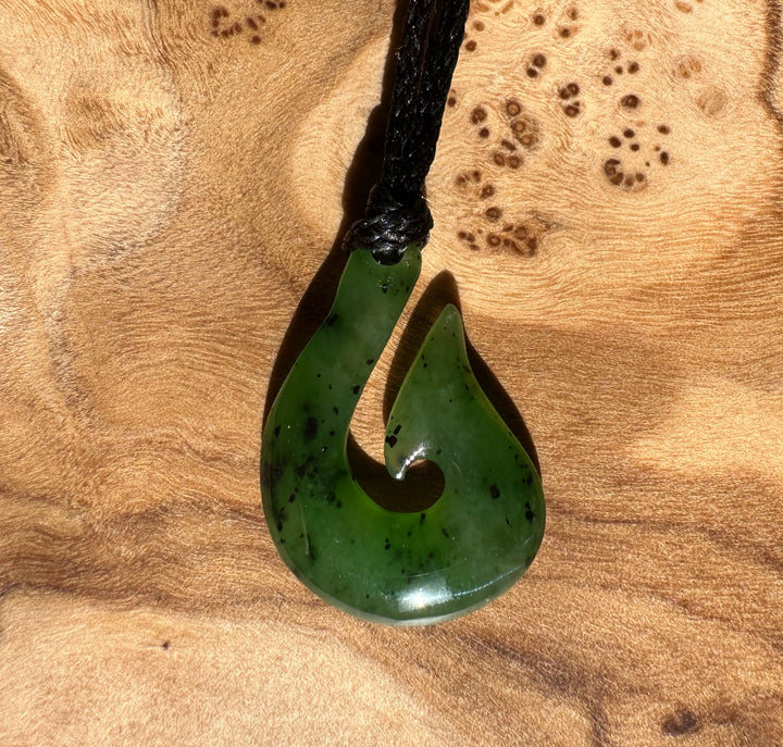 Green jade fish hook pendant on a black cord placed on a warm wood surface with sunlight.