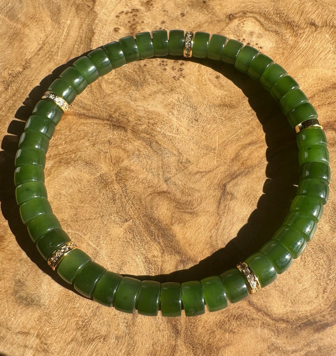 A green jade beaded bracelet with small gold spacer rings, displayed on a textured wooden surface.