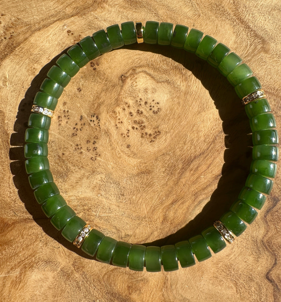 A beaded stretch bracelet made of polished green jade cylindrical beads, accented with small gold spacer rings, displayed on a textured wood surface.
