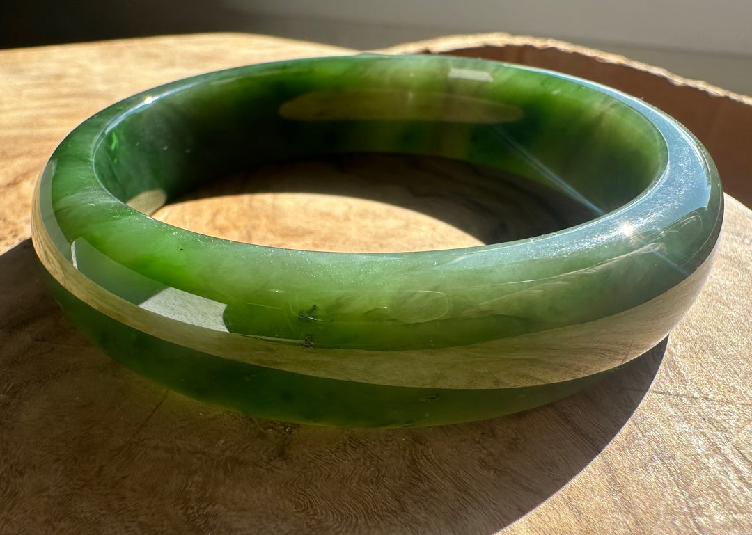 A polished green jade bangle bracelet resting on a wooden surface, showing natural green and white marbled patterns under warm sunlight.