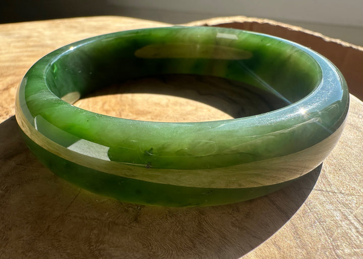 A polished green jade bangle bracelet resting on a wooden surface, showing natural green and white marbled patterns under warm sunlight.