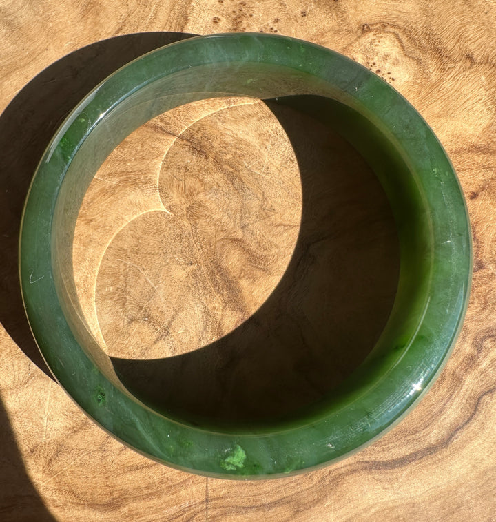 A polished green jade bangle bracelet displayed on a textured wooden surface, casting a distinct shadow.