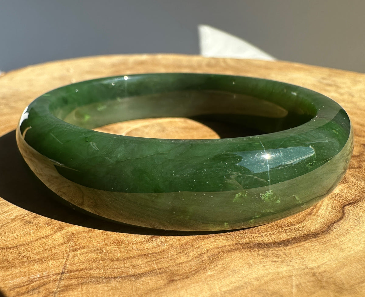A polished green jade bangle bracelet resting on a wooden surface, showing natural green marbling and a glossy finish under direct light.