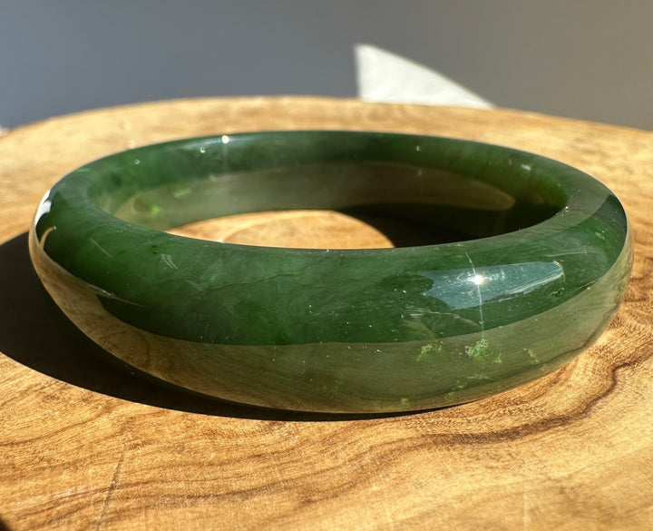A polished green jade bangle bracelet resting on a wooden surface, showing natural green marbling and a glossy finish under direct light.