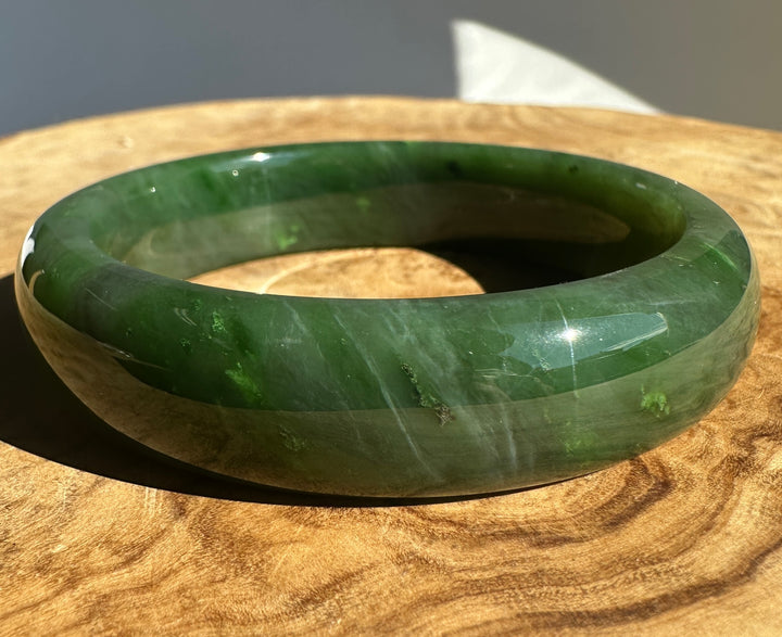 A polished green jade bangle bracelet resting on a round wooden surface under direct sunlight.