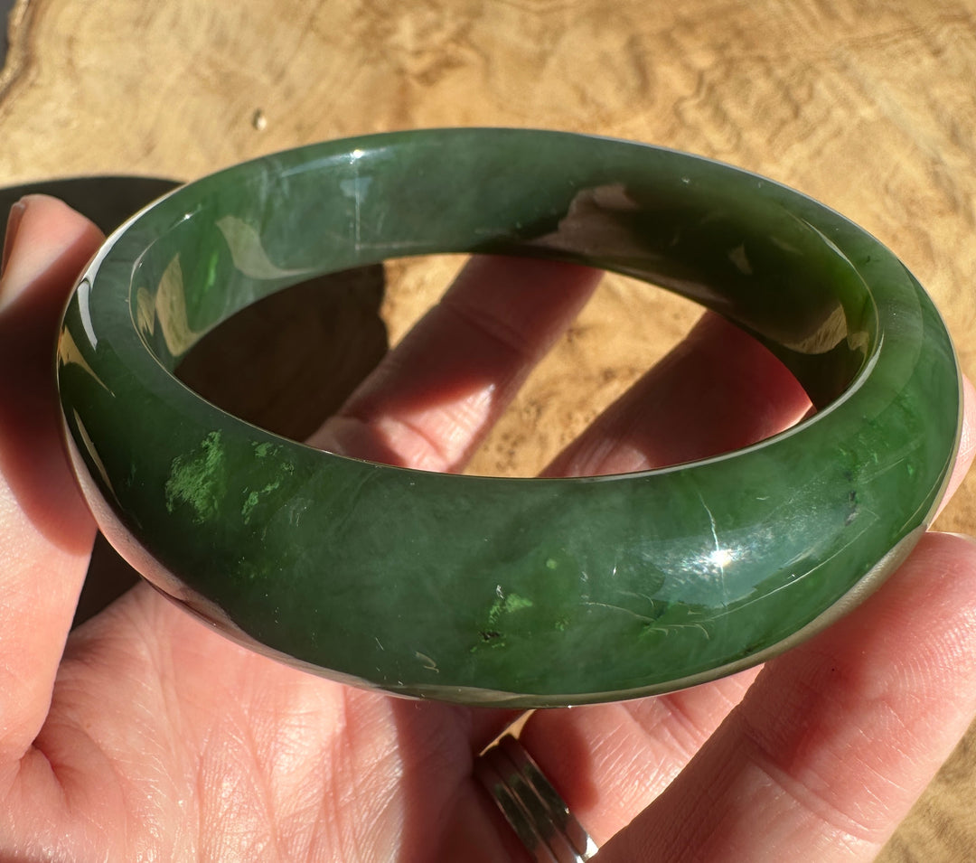 A polished dark green jadeite bangle bracelet held in a hand against a textured tan background.