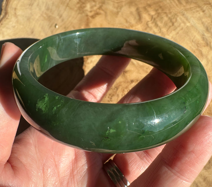 A polished dark green jadeite bangle bracelet held in a hand against a textured tan background.