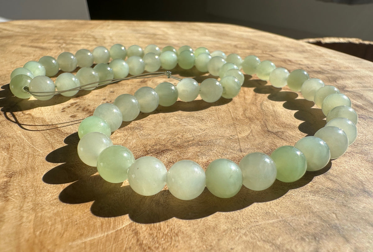A strand of round light green jade-like beads arranged in loops on a natural wooden surface, with sunlight casting shadows.