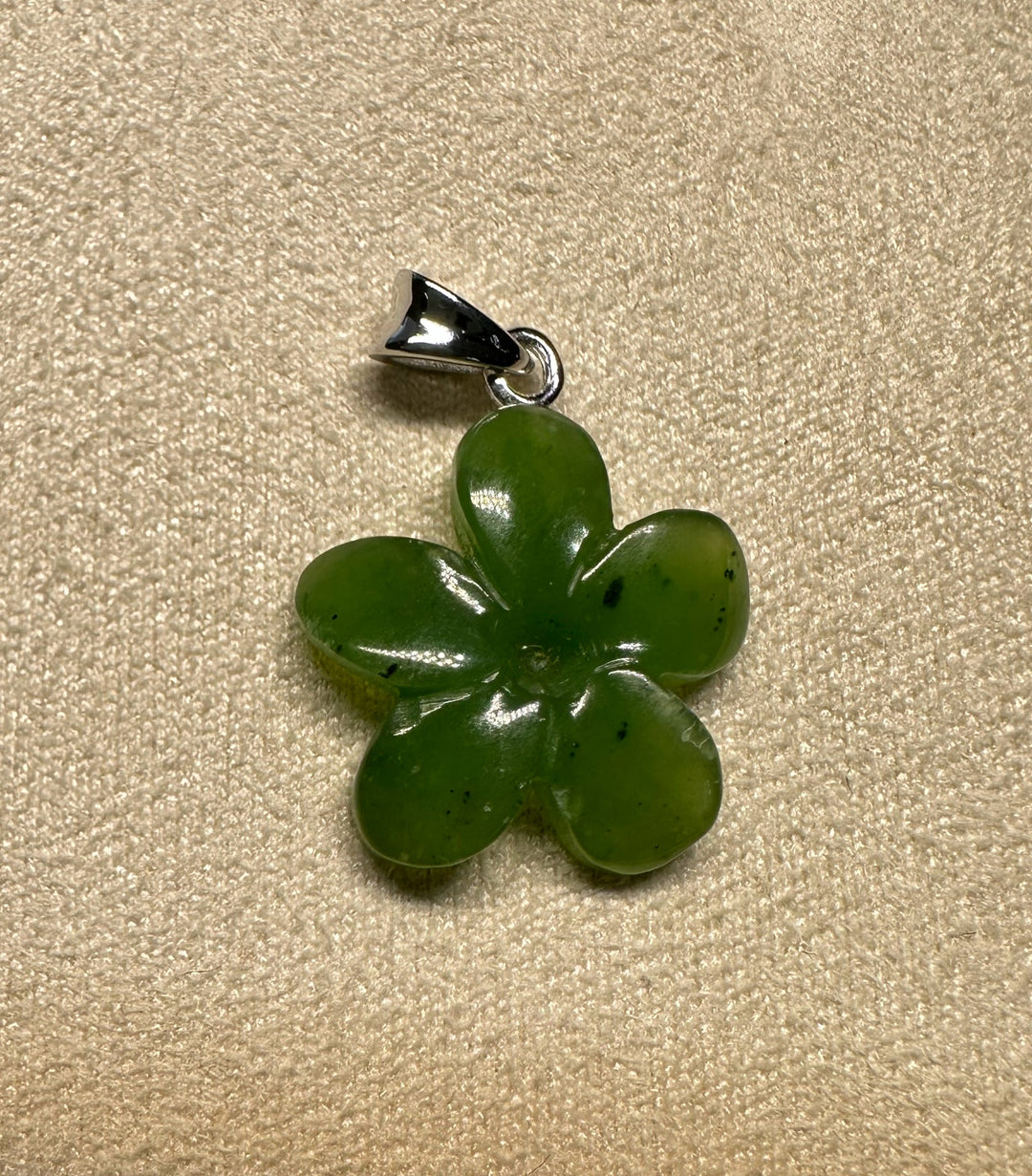 Green jade flower pendant with a silver bail against a textured beige background.
