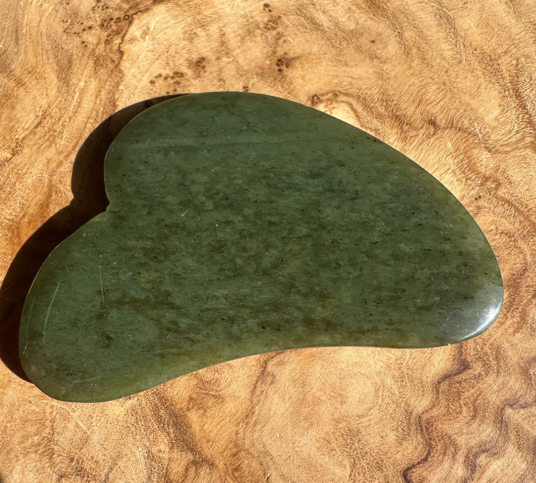 Low grade green nephrite jade gua sha tool with curved edge on wooden surface