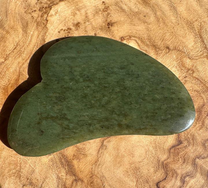 Low grade green nephrite jade gua sha tool with curved edge on wooden surface