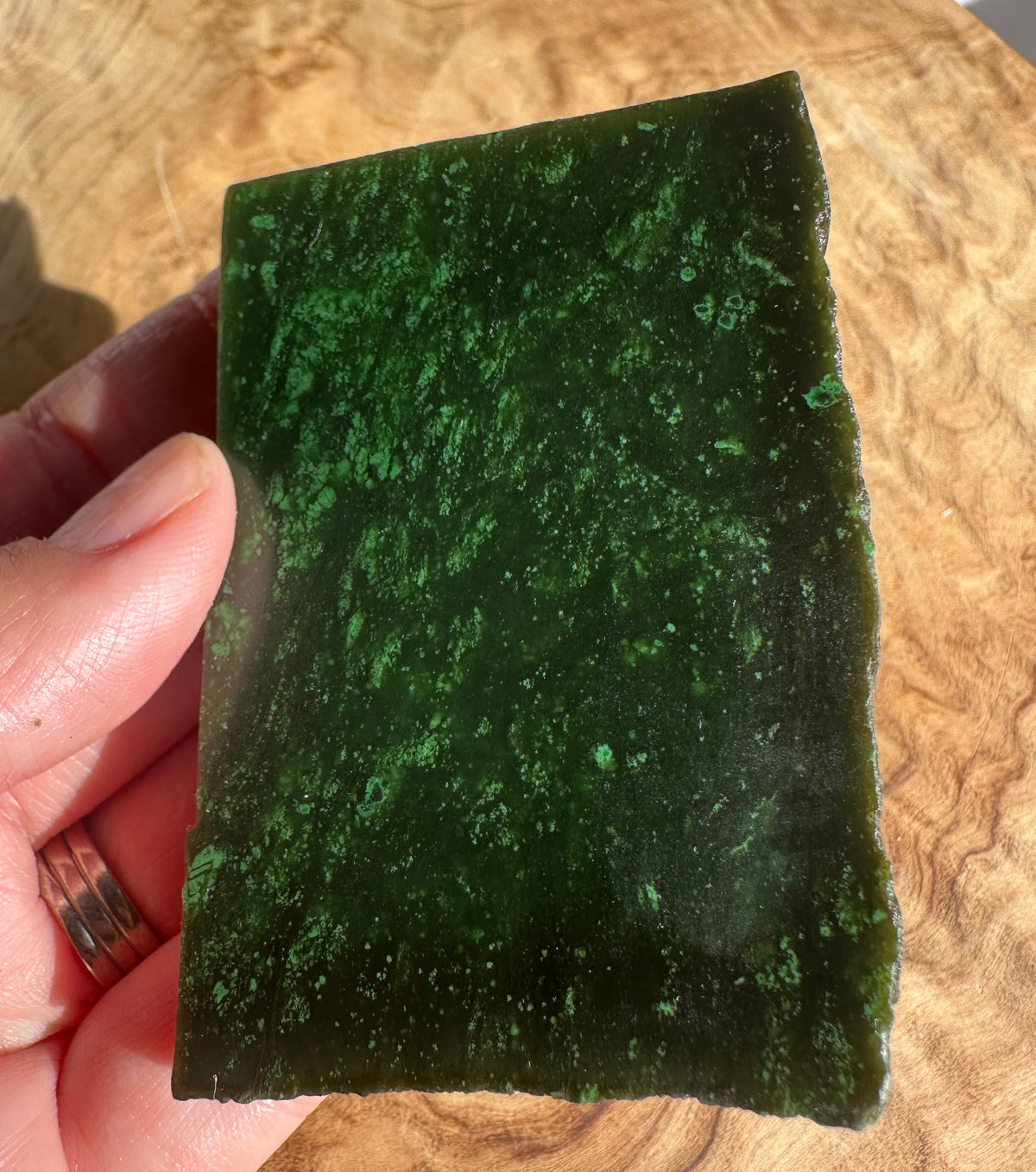 Cassiar Jade Slab 60grams dark green with lighter green speckles held by fingers over a wood surface