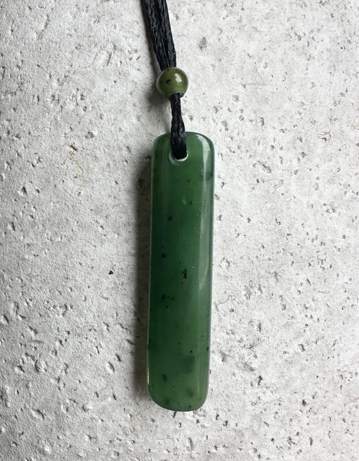 Green jade drop pendant on a black cord, with a jade bead on top. Grey cement background