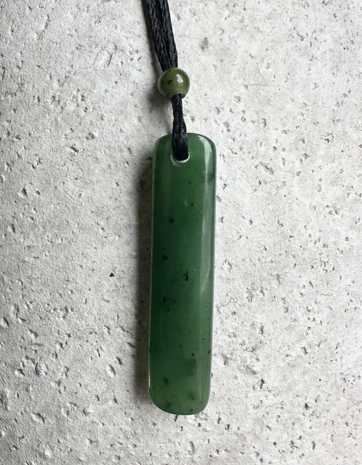 Green jade drop pendant on a black cord, with a jade bead on top. Grey cement background