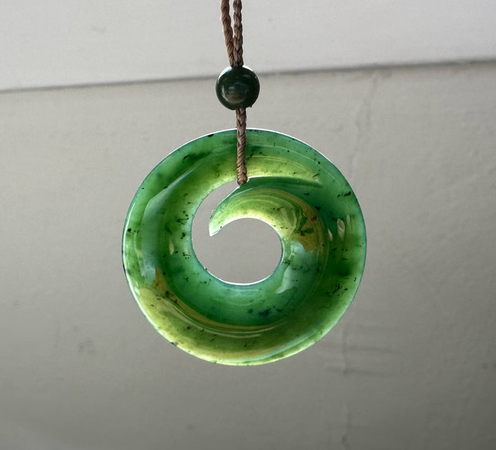 Circular green jade pendant with a large hole hanging from a brown braided cord with a small green bead above