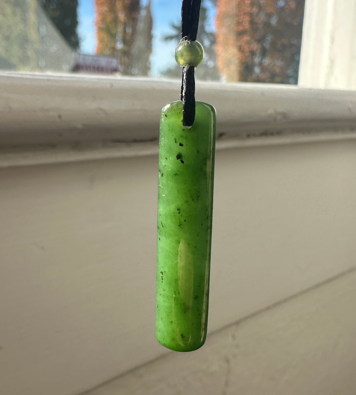 Pendant Drop 2054-1 #4 in green stone with a round bead above, hung on a black cord against a sunlit wooden background