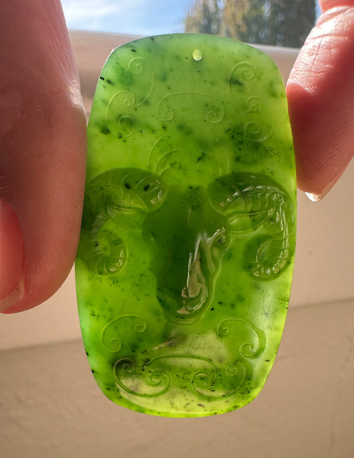 Bright green Canadian jade ram pendant with curling scroll patterns held between fingers.