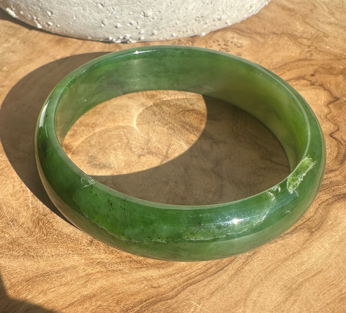 A round green jade bangle bracelet resting on a wooden surface with a shadow cast to the left.