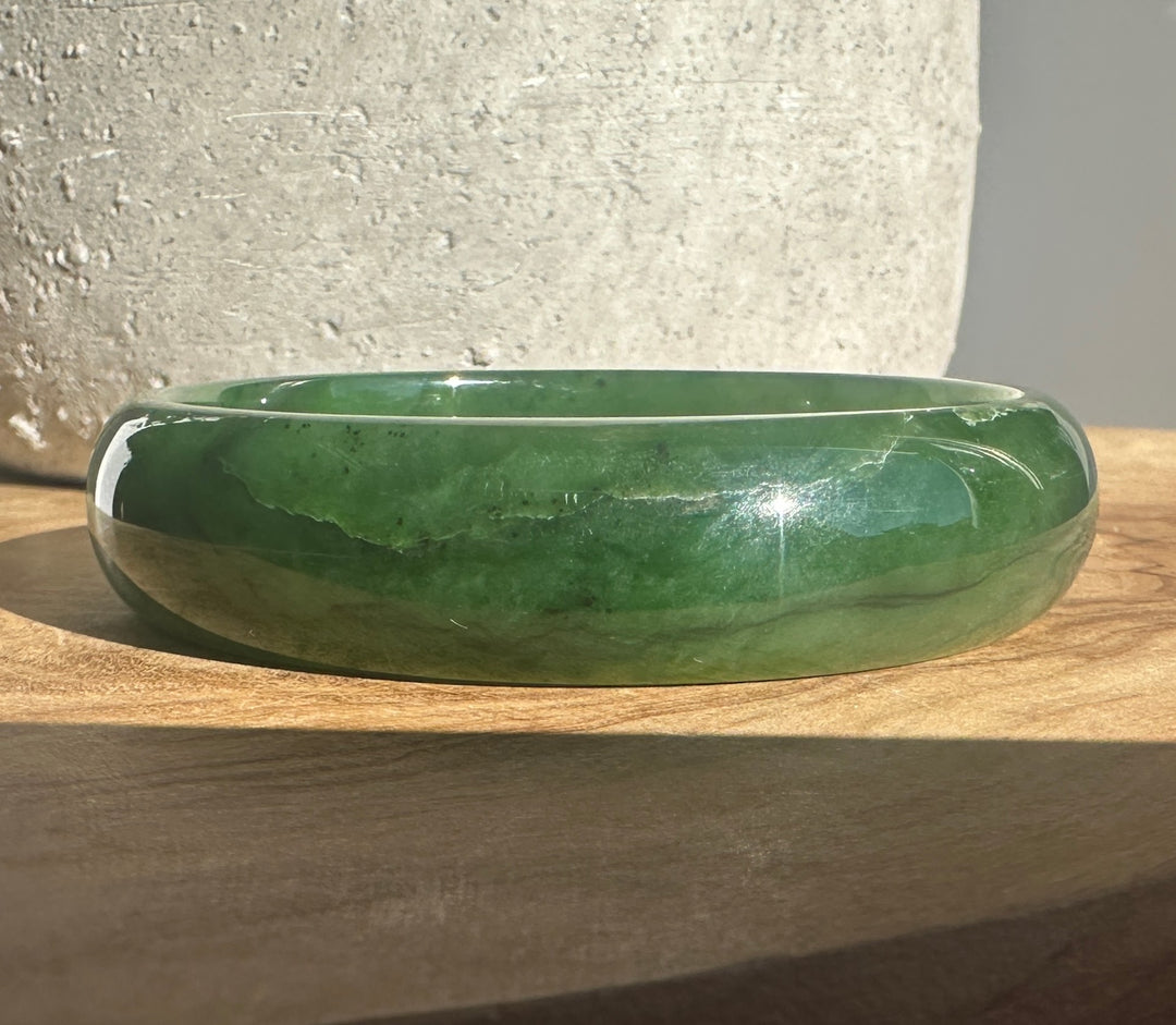 A polished green jade bangle bracelet resting on a wooden surface with a textured gray ceramic pot in the background.