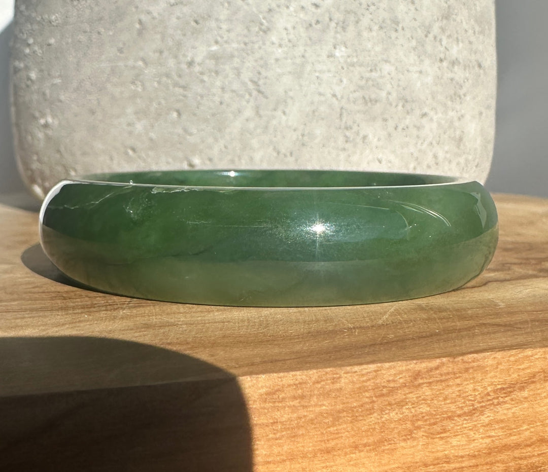 A polished green jade bangle bracelet resting on a wooden surface with a textured stone vase in the background.