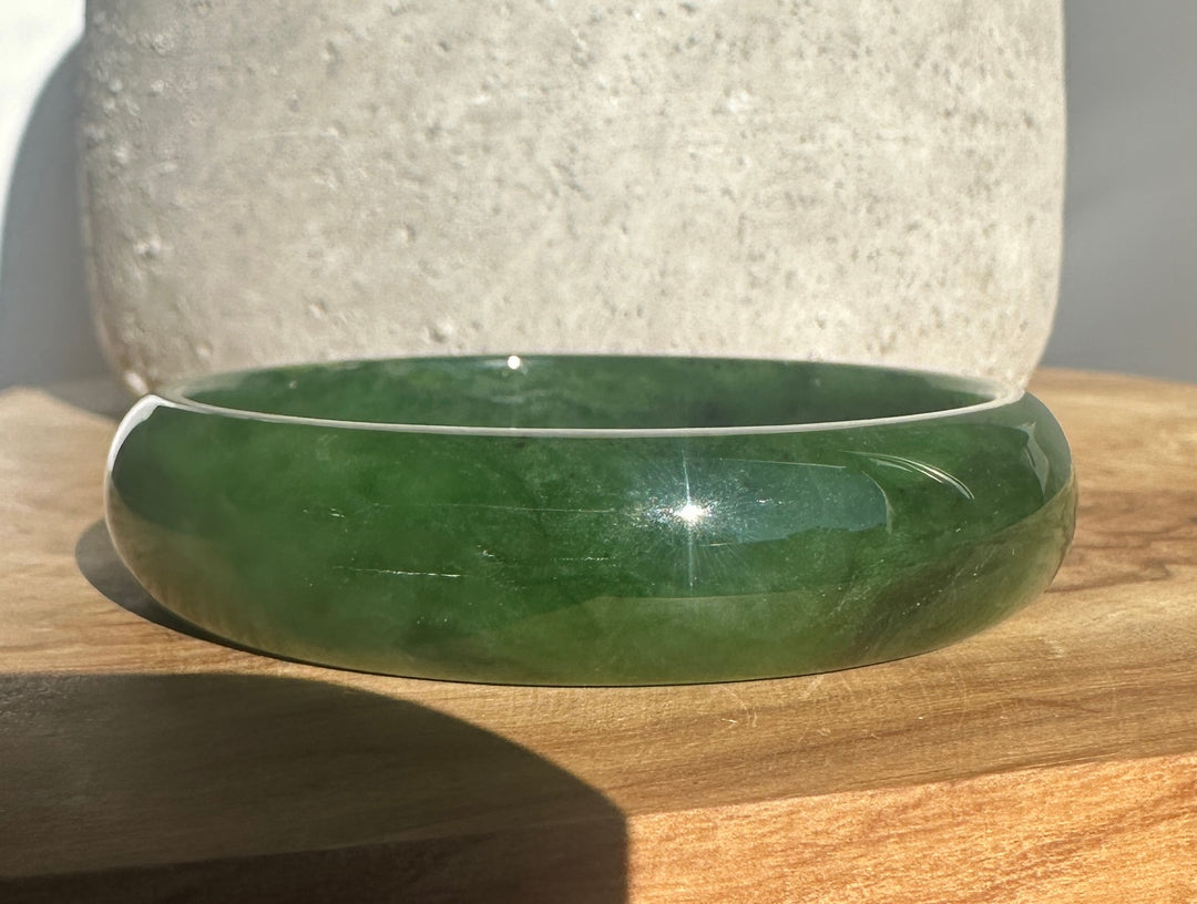A green jade bangle bracelet with natural color variations, resting on a wooden surface.
