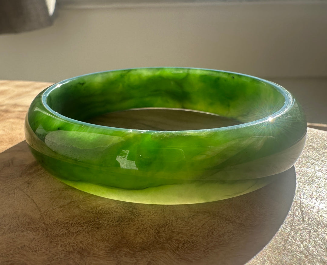A round green jadeite bangle bracelet with natural marbled patterns, resting on a wooden surface under direct sunlight.