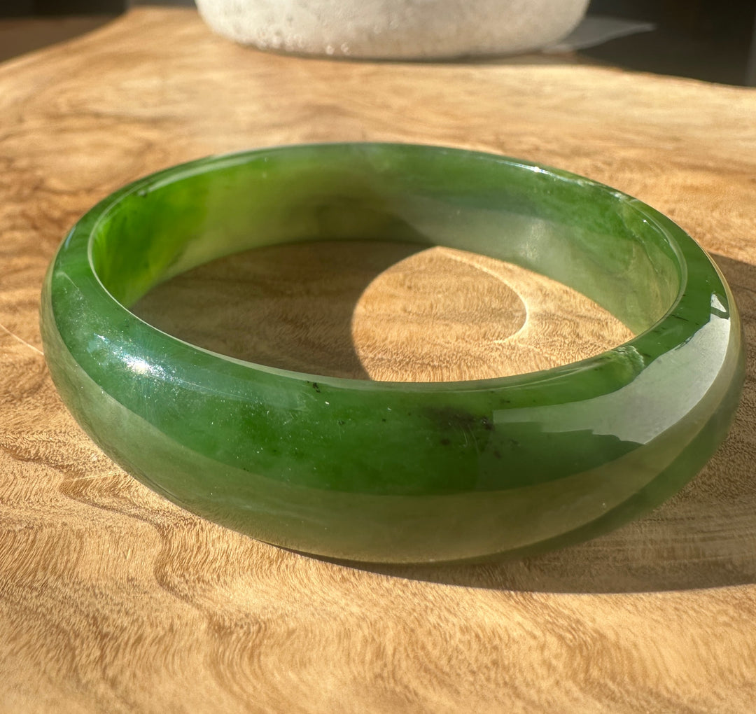A green jade bangle bracelet with natural color variations, resting on a wooden surface.